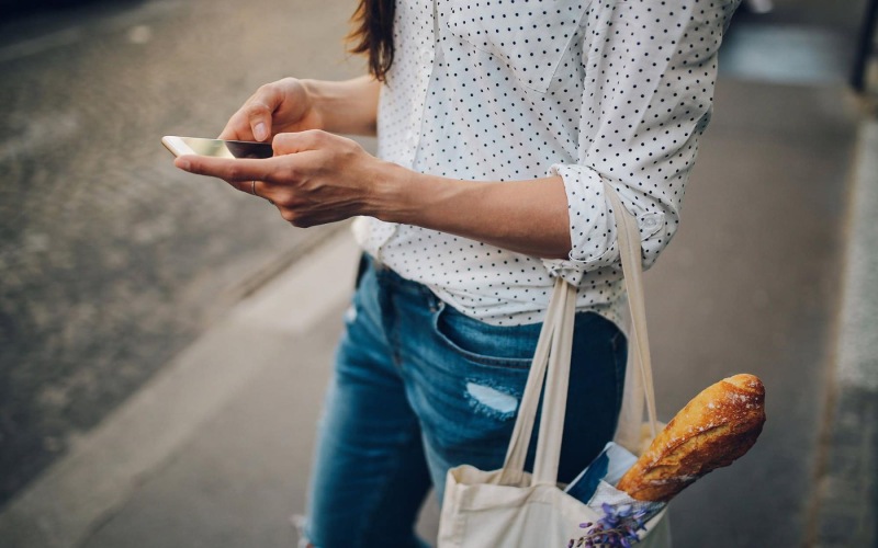 woman walks down the street and texts with a bag of groceries on her arm