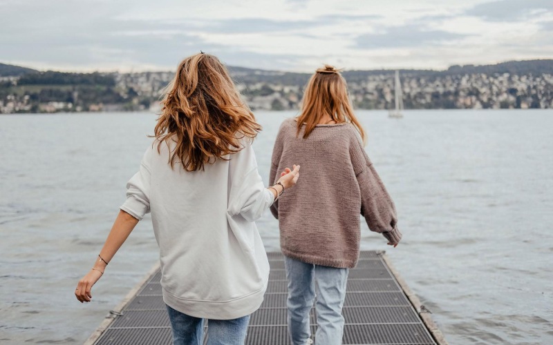 two women walk to the edge of a dock, in a body of water, looking toward the city on the other shore