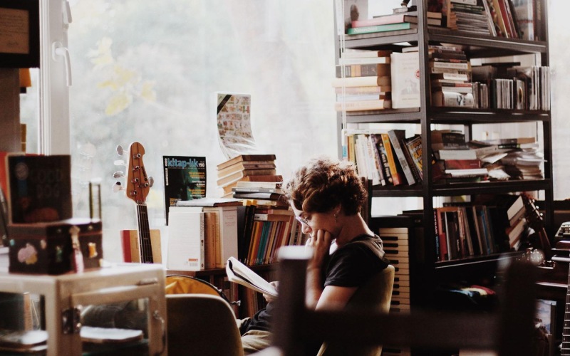 woman, surrounded by books, sits and reads in front of a picture window