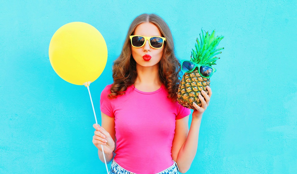 woman against a blue background, holding a pineapple in one hand and balloon in the other
