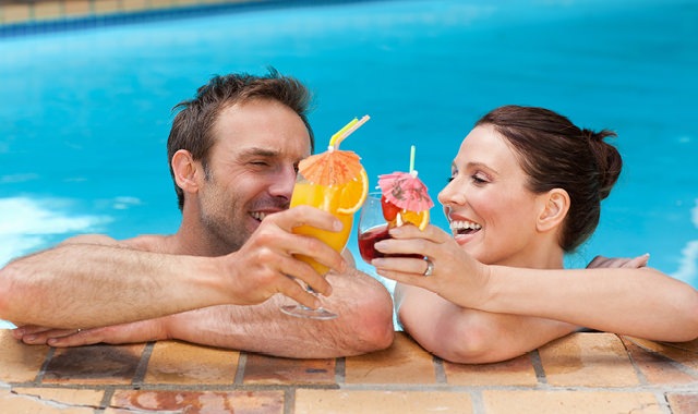 happy couple in the pool with arms resting on the ledge, drinks in hand, as they toast each other