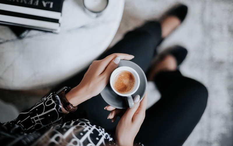 business woman sits next to a marble table with books and holds a saucer with a cup of espresso
