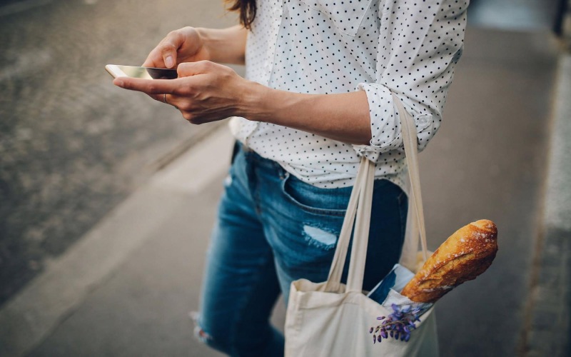 woman walks down the street and texts with a bag of groceries on her arm